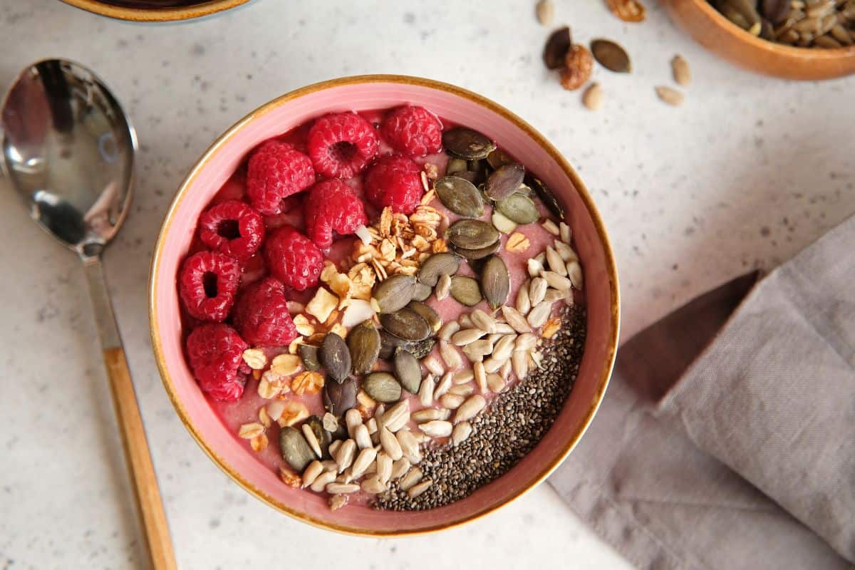 Pink smoothie bowl topped with raspberries, chia seeds, sunflower seeds, and granola.