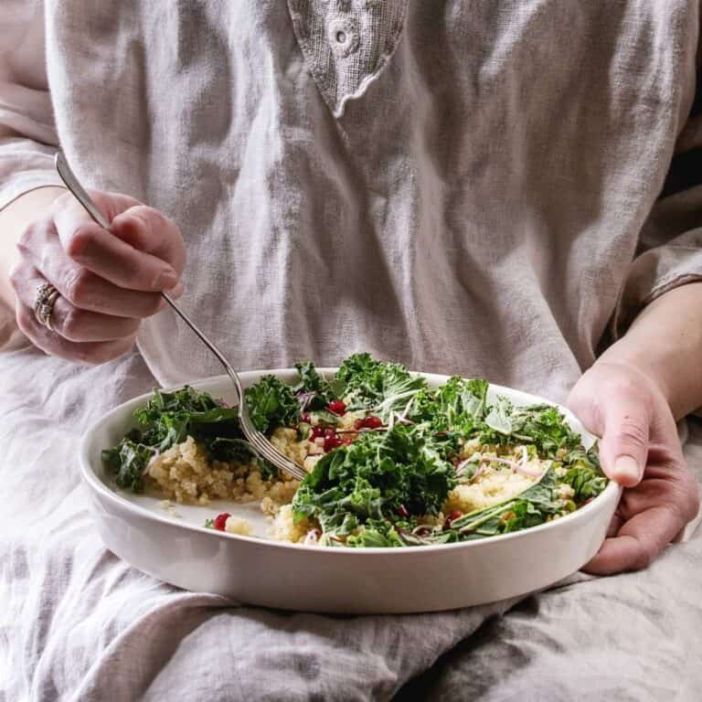Person holding a bowl of quinoa and leafy green salad topped with kale and pomegranate seeds.