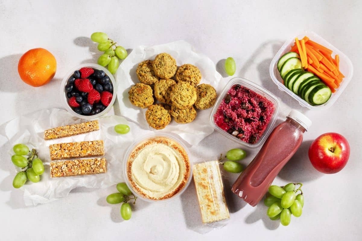 Assortment of healthy vegan snacks including hummus, fresh fruit, vegetables, crackers, and a smoothie arranged on a table.