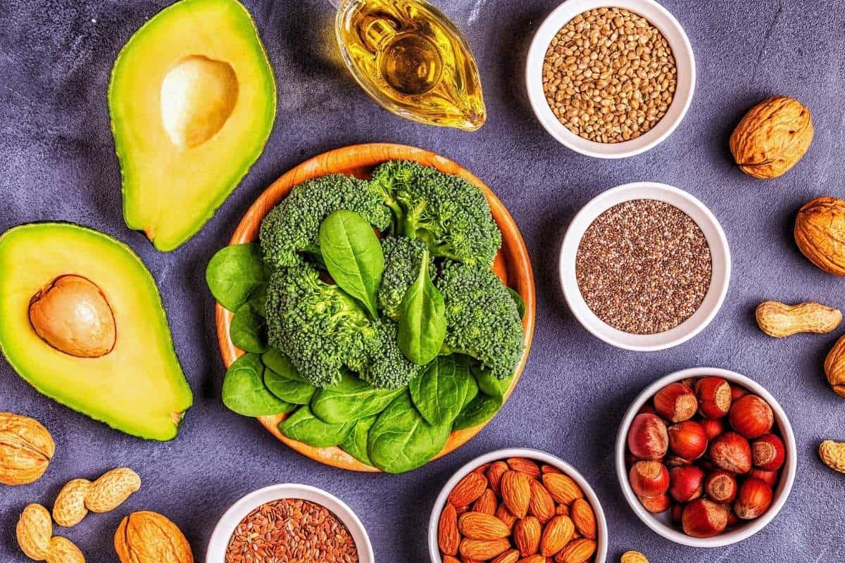 Plant-based foods including avocado, broccoli, spinach, nuts, seeds, and grains arranged on a dark background.