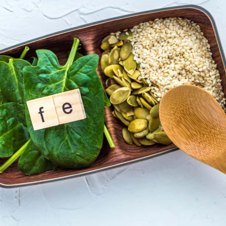 Spinach leaves, pumpkin seeds, and sesame seeds arranged on a tray with letter tiles spelling “Fe” for iron.