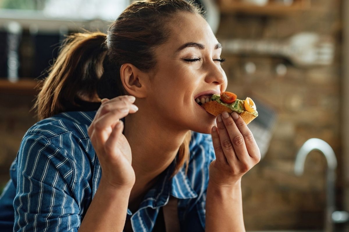 A woman in a blue plaid shirt enjoying a bite of food in a kitchen.