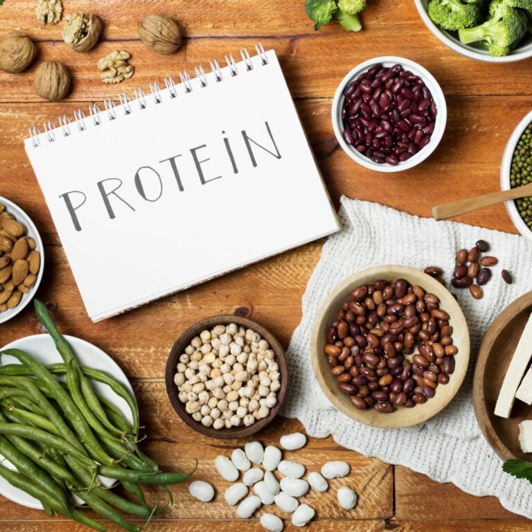 An open notebook labeled “PROTEIN” surrounded by bowls of beans, lentils, nuts, chickpeas, broccoli, and other plant-based protein sources on a wooden table.