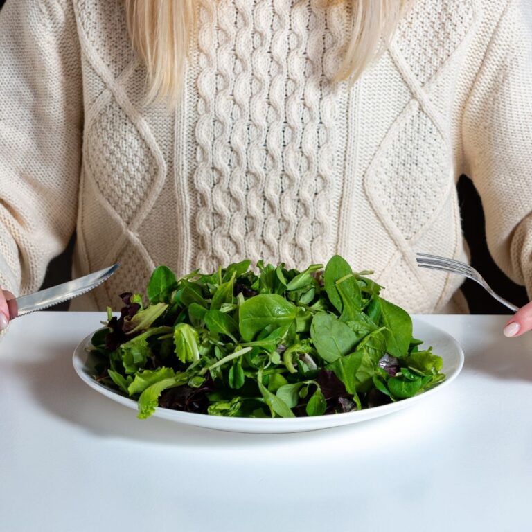 Woman in a white knitted sweater sitting at a table with a plate of fresh leafy green salad, holding a knife and fork.