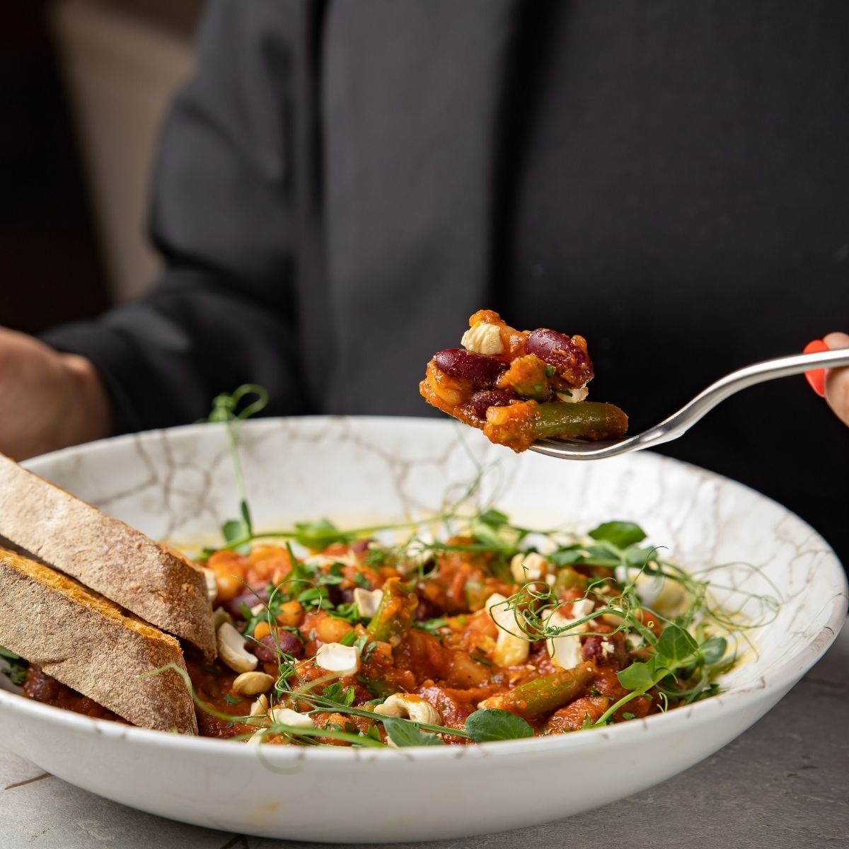 Person holding a spoonful of vegetable and bean stew served in a white bowl with fresh greens and slices of bread.