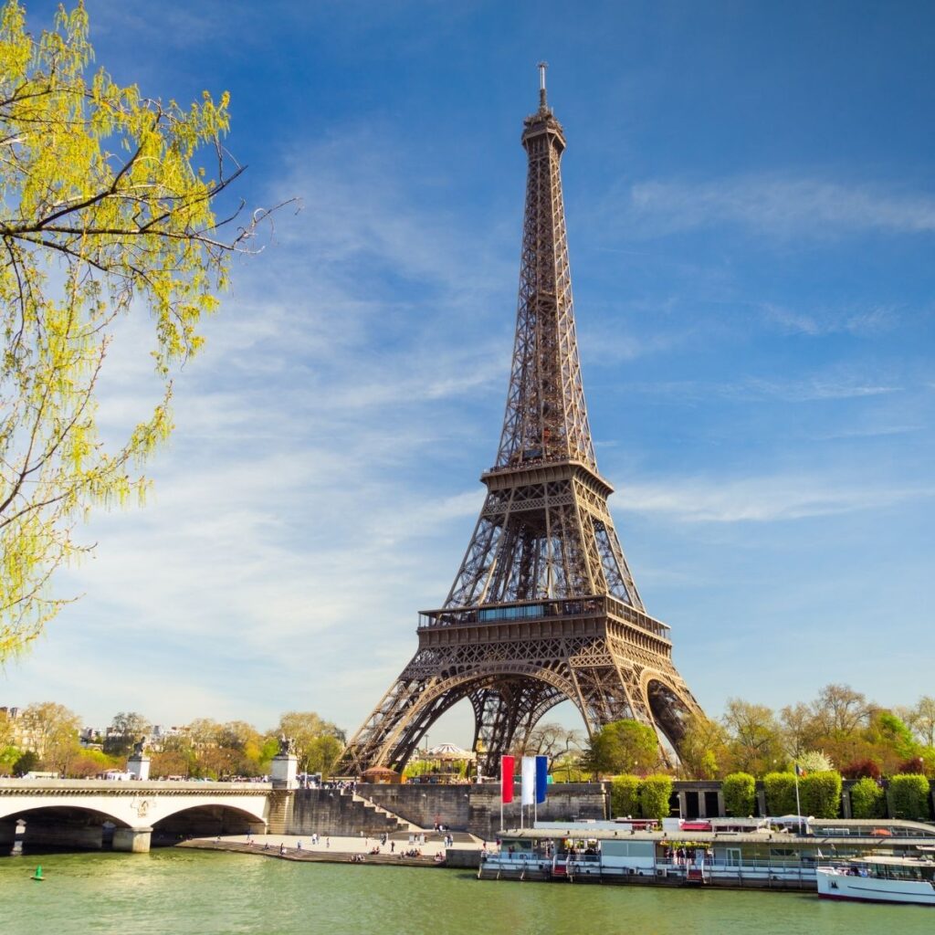 The Eiffel Tower in Paris, France, overlooking the Seine River with a stone bridge, green trees, and a clear blue sky.
