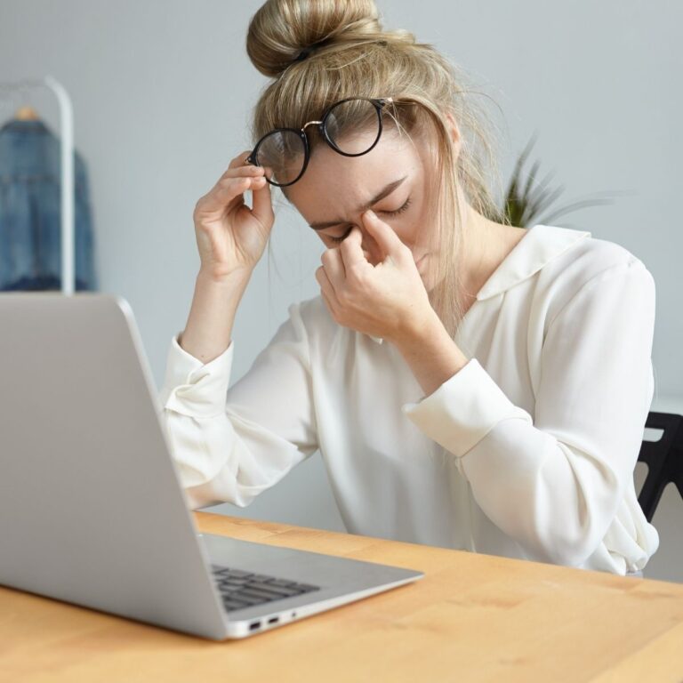 Tired woman sitting at a laptop, rubbing her eyes and pinching the bridge of her nose while holding her glasses.