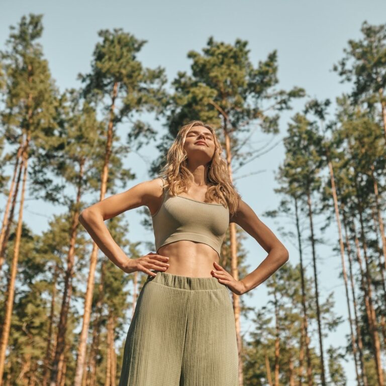Smiling woman standing outdoors in a pine forest with her hands on her hips, wearing green workout clothes and enjoying the sunshine.