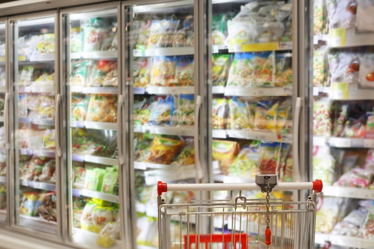 Shopping cart in front of refrigerated shelves filled with packaged foods at a grocery store.