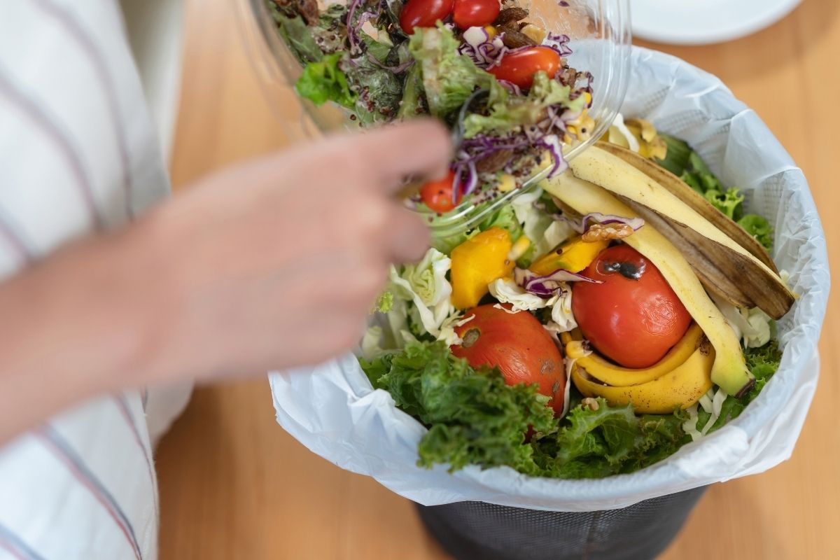 Person throwing fresh vegetables and food scraps into a kitchen waste bin.