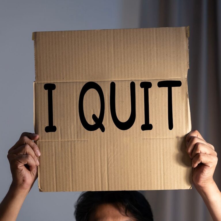 Person holding a cardboard sign with the words "I QUIT" written in large black letters, covering their face.