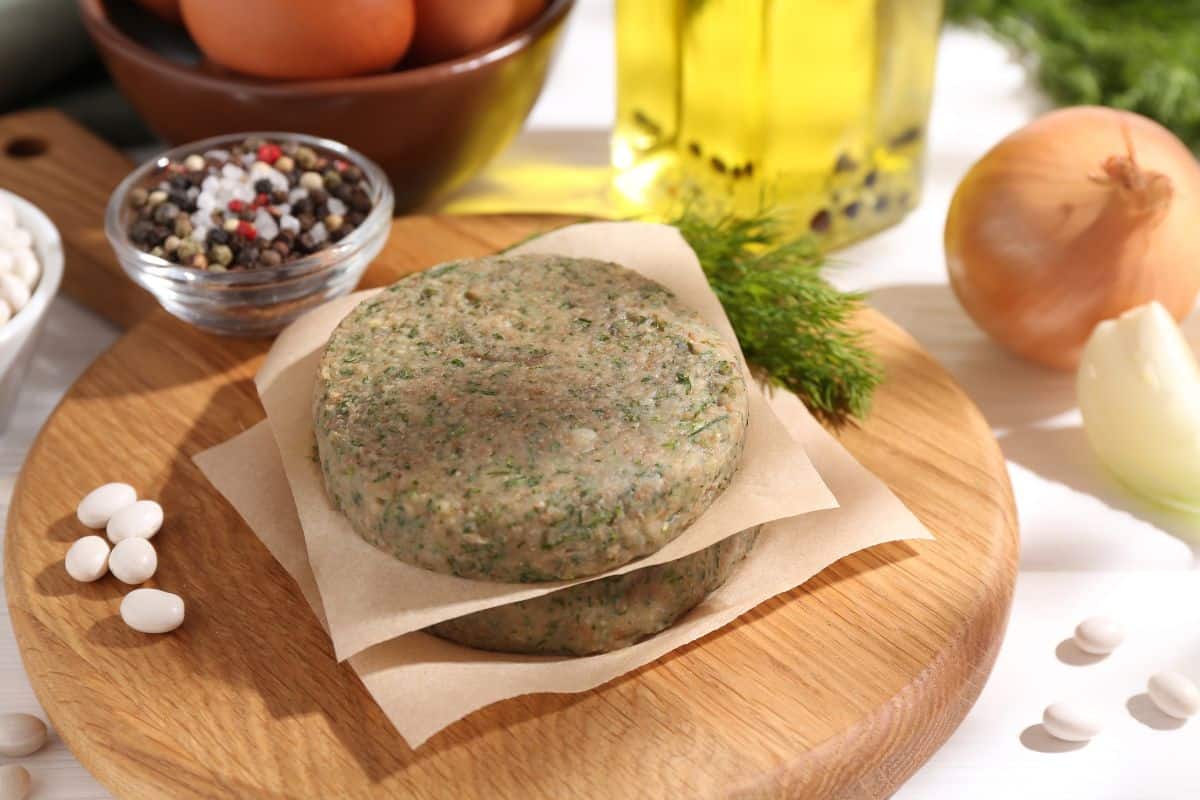 Uncooked plant-based burger patties on parchment with herbs, spices, and vegetables in the background.