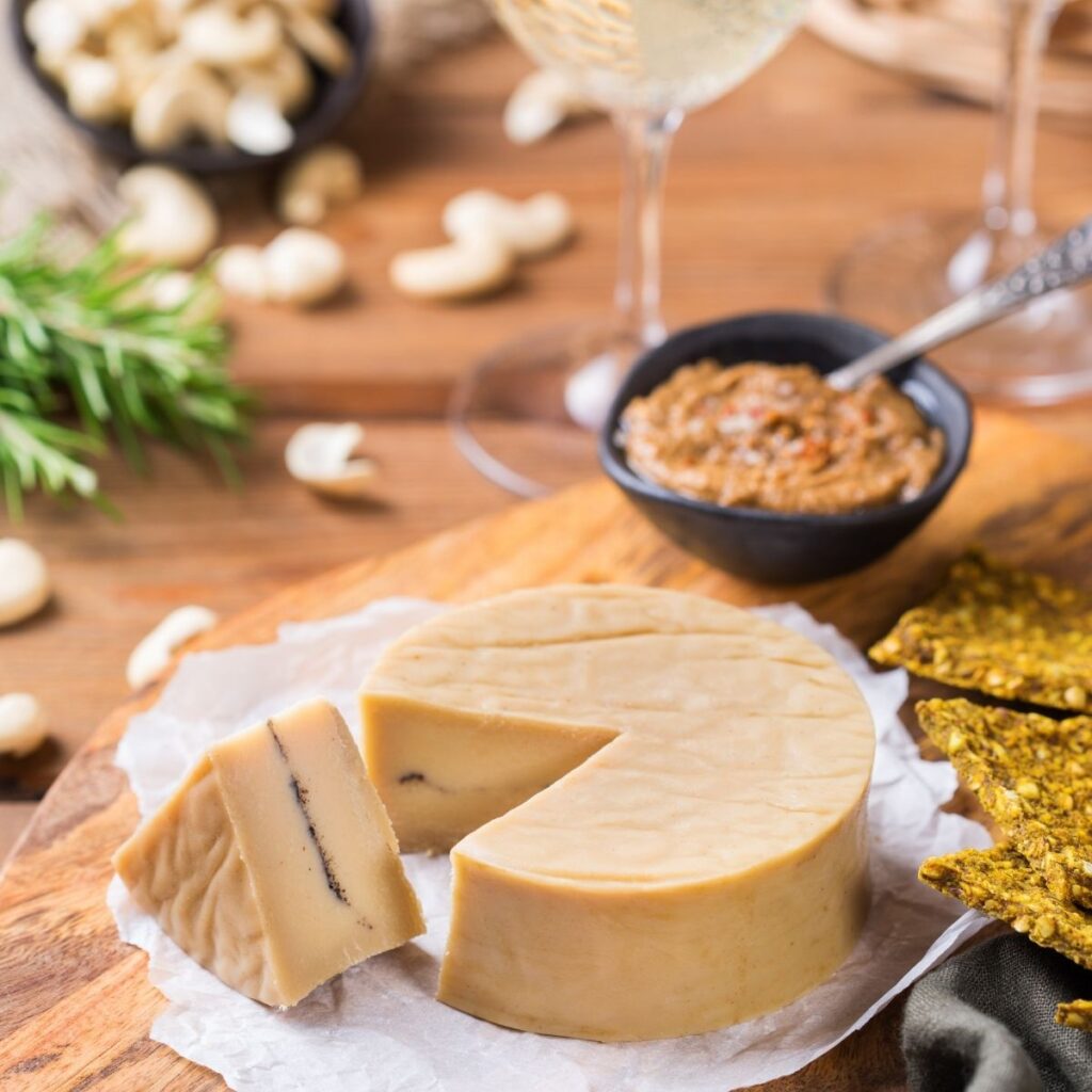 Round wheel of vegan cheese with a sliced wedge on a wooden board, served with crackers and cashews.
