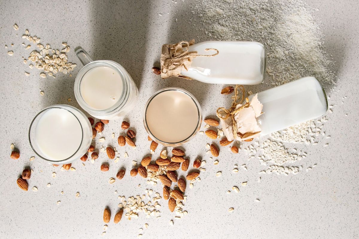 Assorted plant-based milks in glasses and bottles with oats and almonds on a light surface.