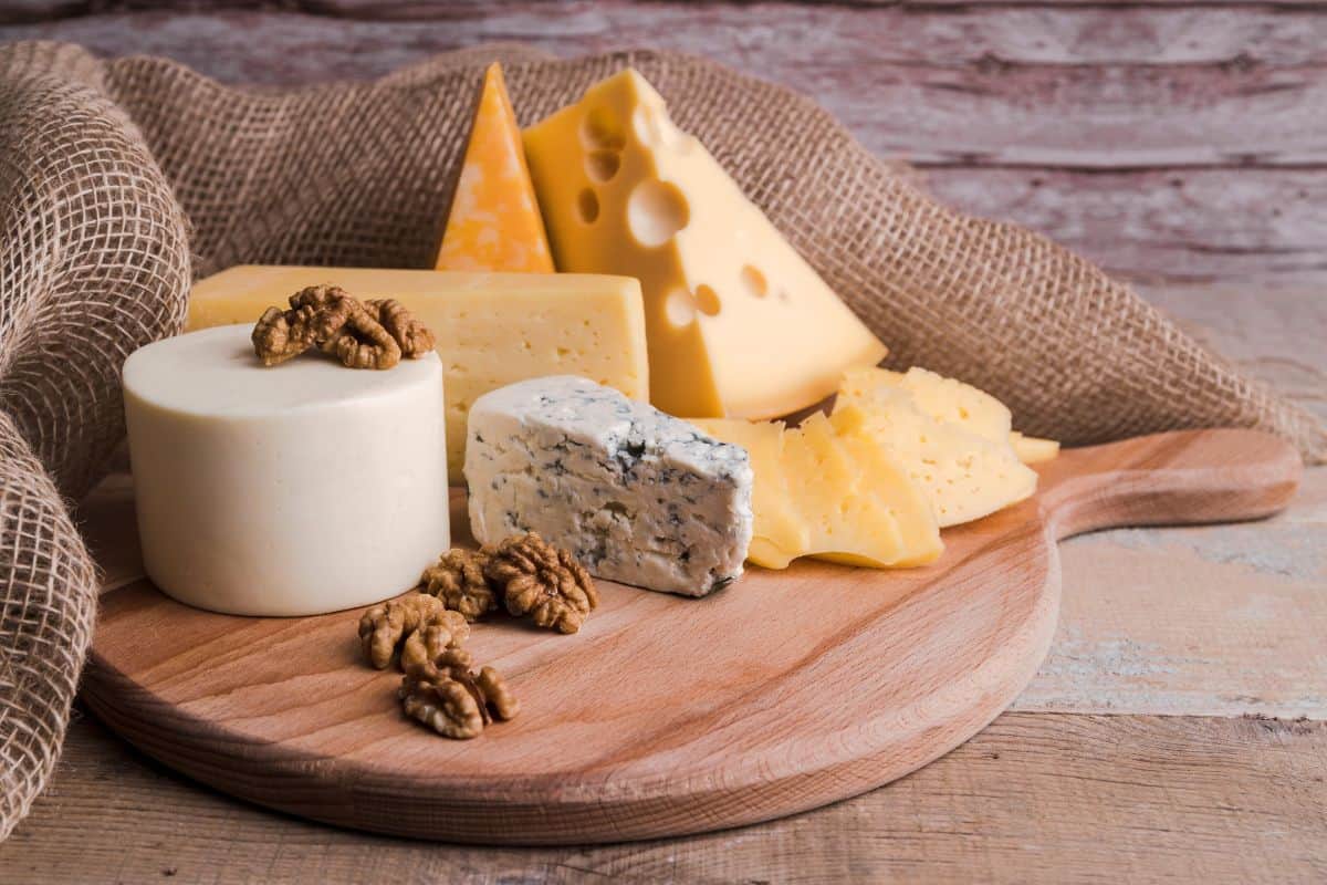 Assorted cheeses, including blue cheese and sliced hard cheeses, arranged on a wooden board with walnuts.