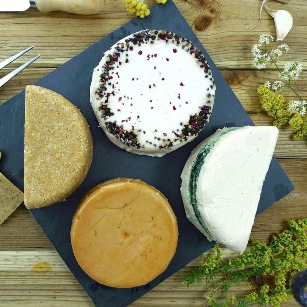 Four varieties of artisanal vegan cheeses on a slate board with assorted crackers, surrounded by rustic flowers and a cheese knife