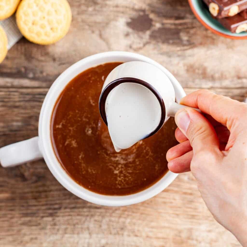 A hand pours cream into a cup of coffee on a wooden table. Nearby are cookies on a napkin and a bowl of chocolate