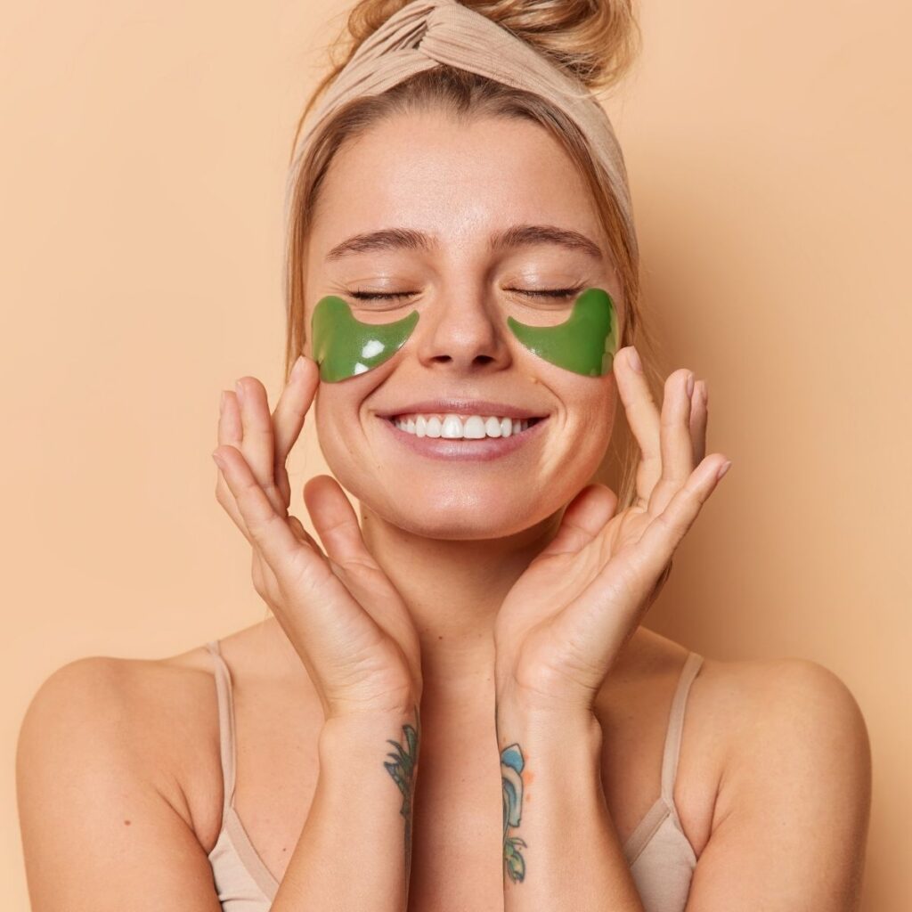Woman smiling while wearing green under-eye skincare patches against a warm beige background.