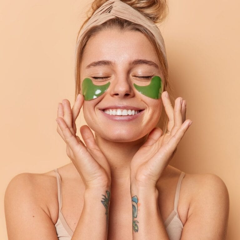 Woman smiling while wearing green under-eye skincare patches against a warm beige background.