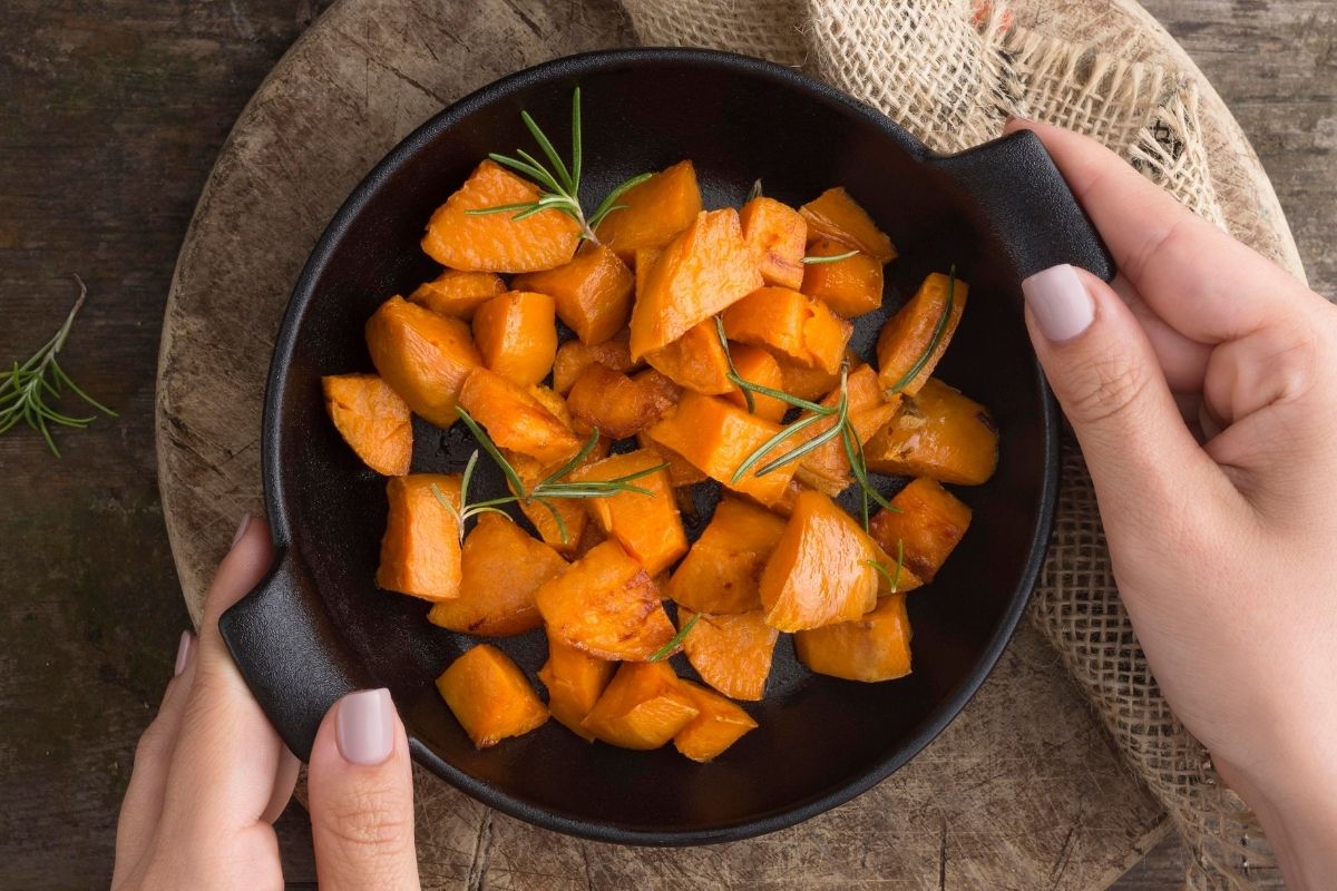 Overhead view of roasted orange sweet potato cubes garnished with fresh rosemary in a black bowl held by two hands.