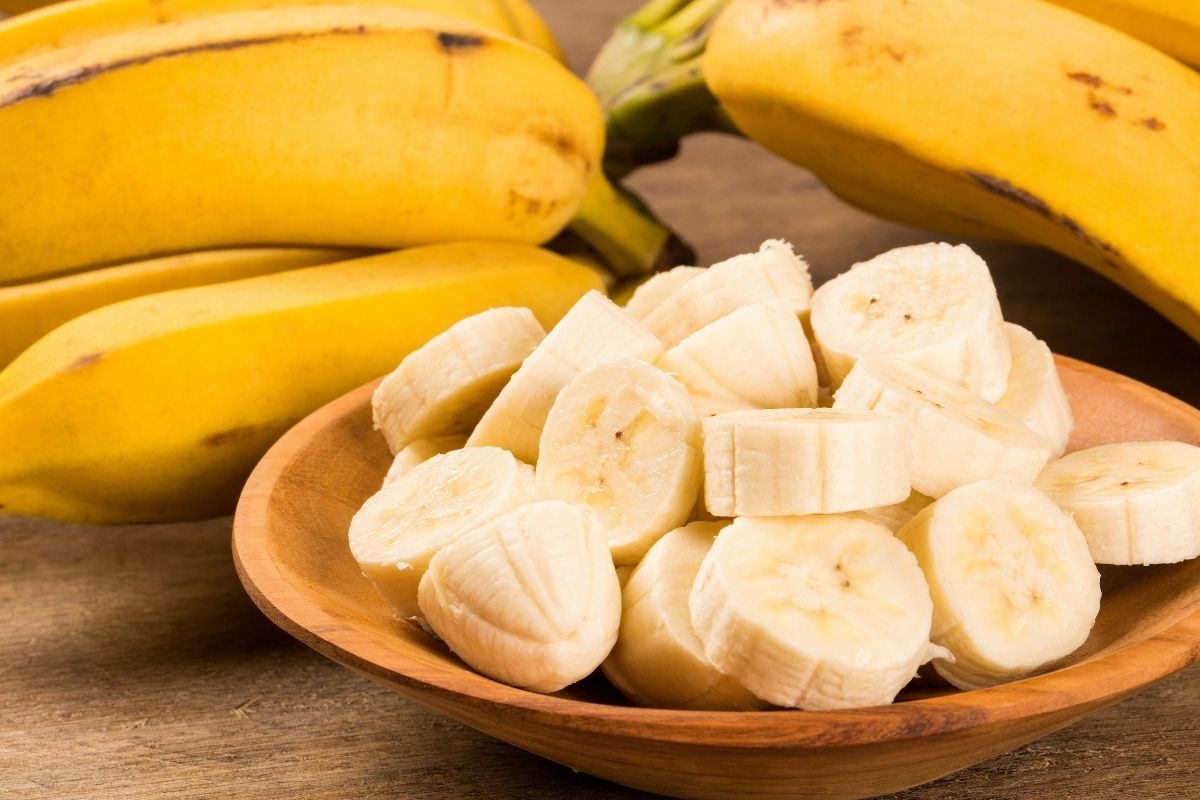 Wooden bowl filled with sliced bananas, with bunches of ripe yellow bananas in the background on a rustic table.
