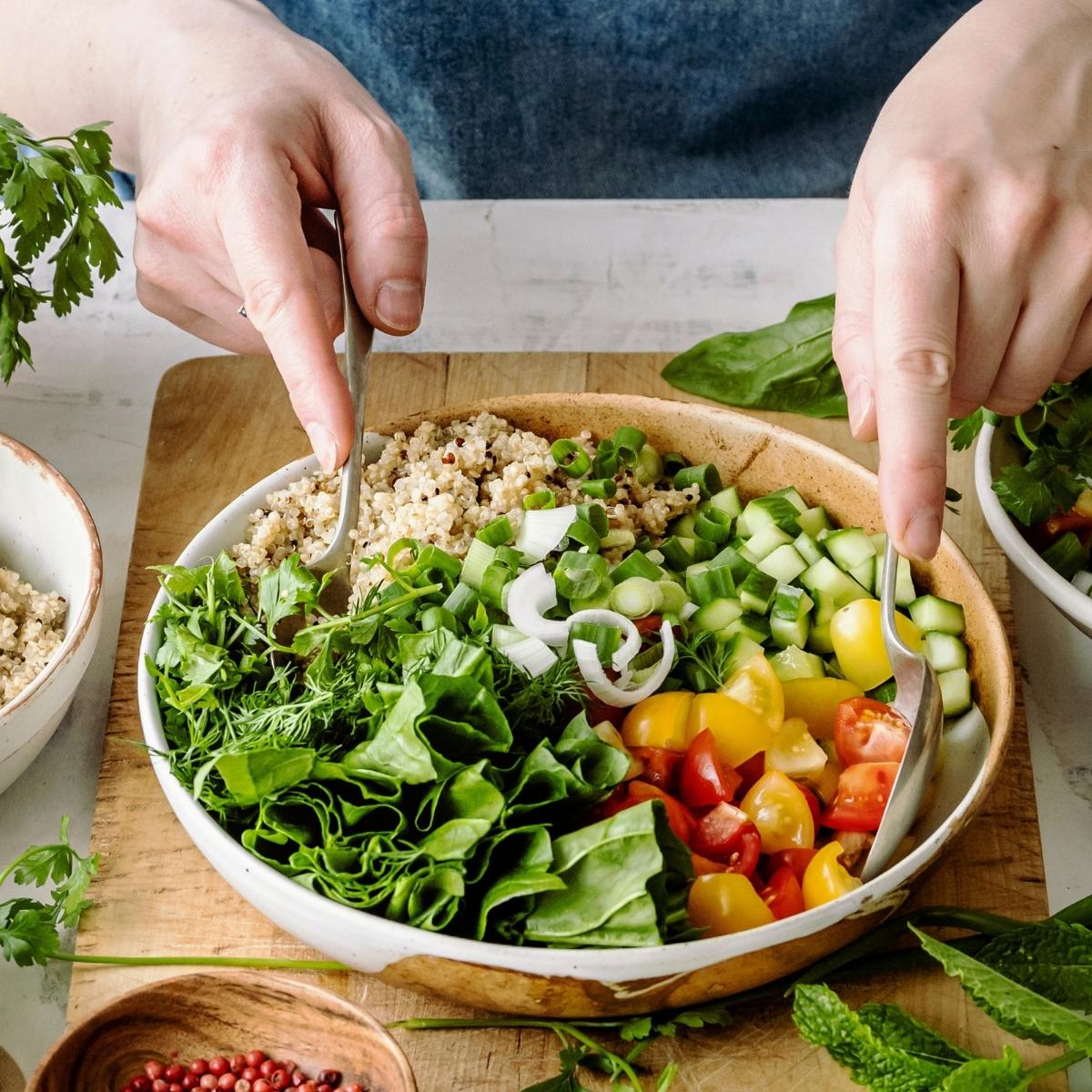 Person mixing a colorful vegan salad with leafy greens, quinoa, cucumber, cherry tomatoes, and green onions in a large bowl.