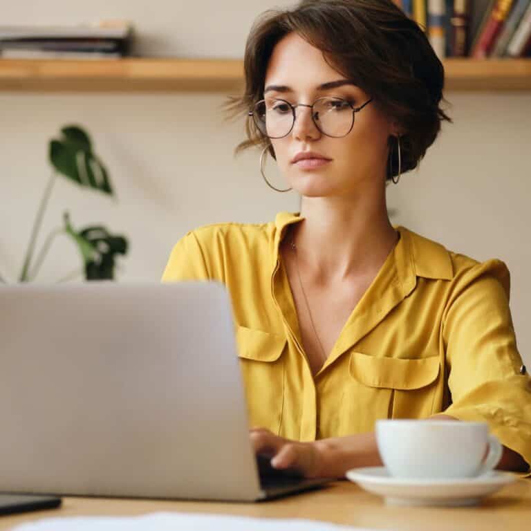 Woman wearing glasses working on a laptop at home with a cup of coffee beside her.