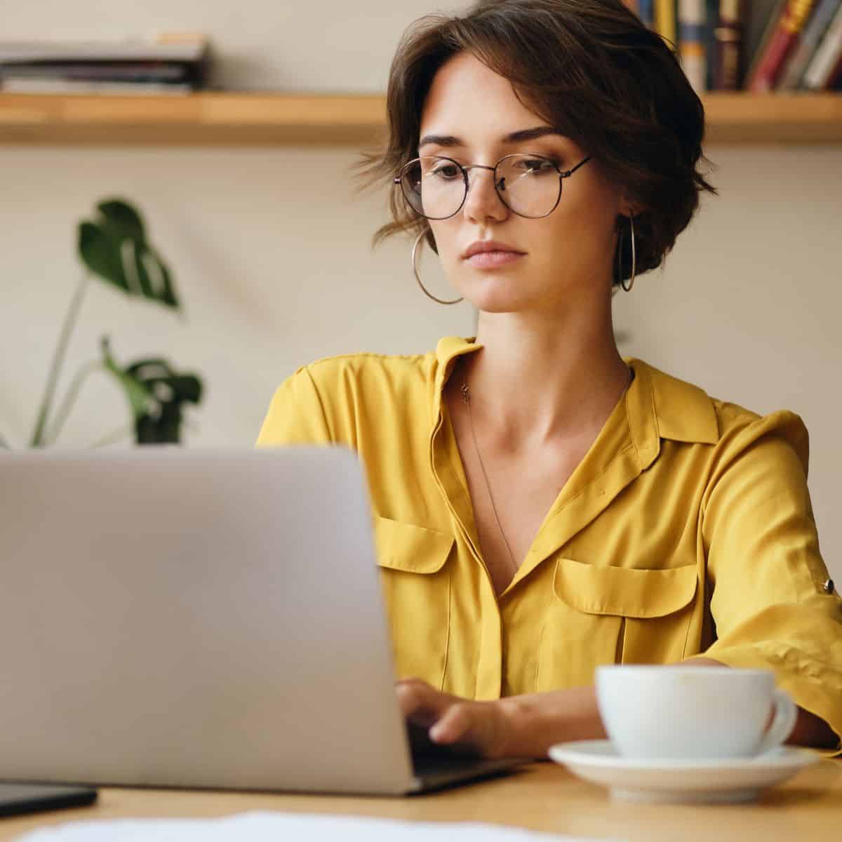 Woman wearing glasses working on a laptop at home with a cup of coffee beside her.