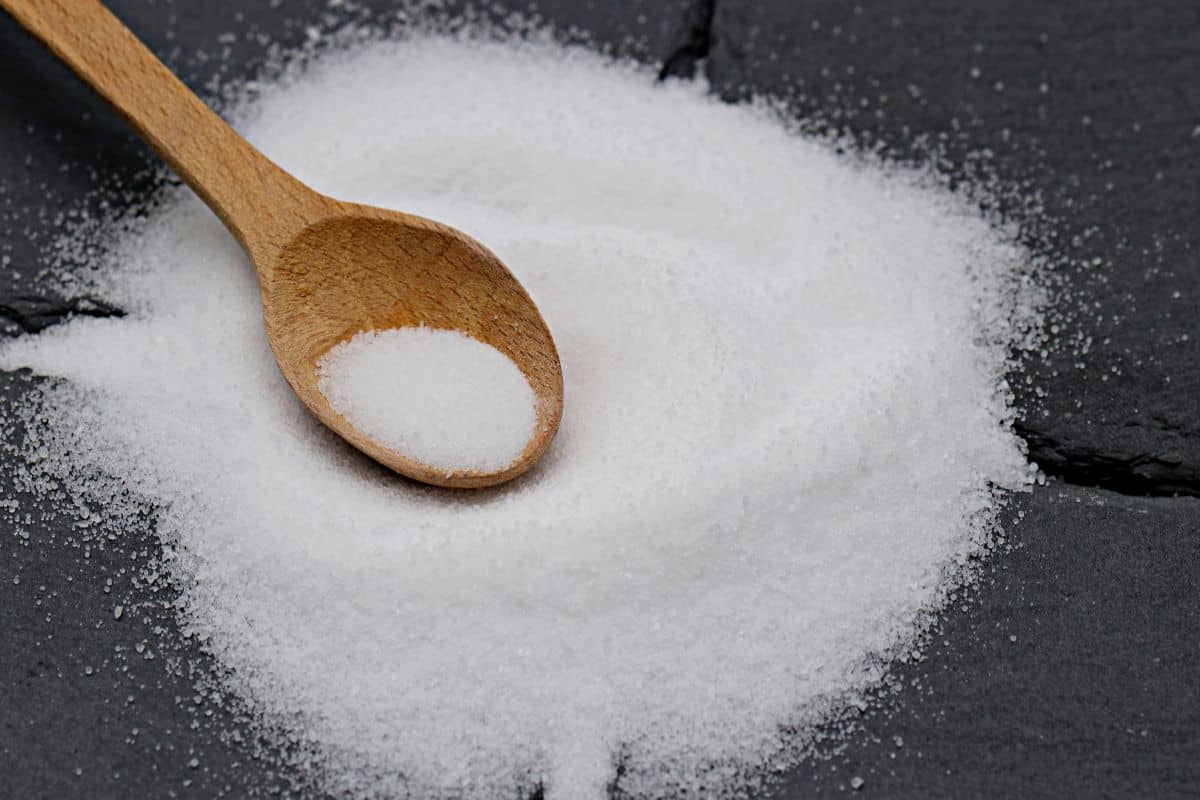 Granulated white sugar spread on a dark surface with a wooden spoon resting on top.
