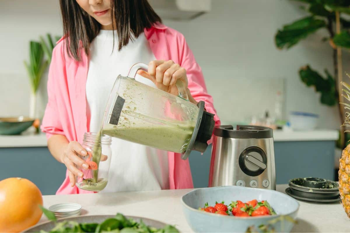 Woman pouring a plant-based smoothie from a blender into a glass in a bright kitchen.