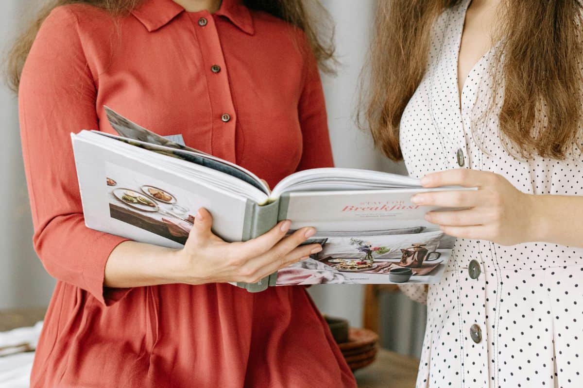 Two women looking at a cookbook while preparing food in kitchen.