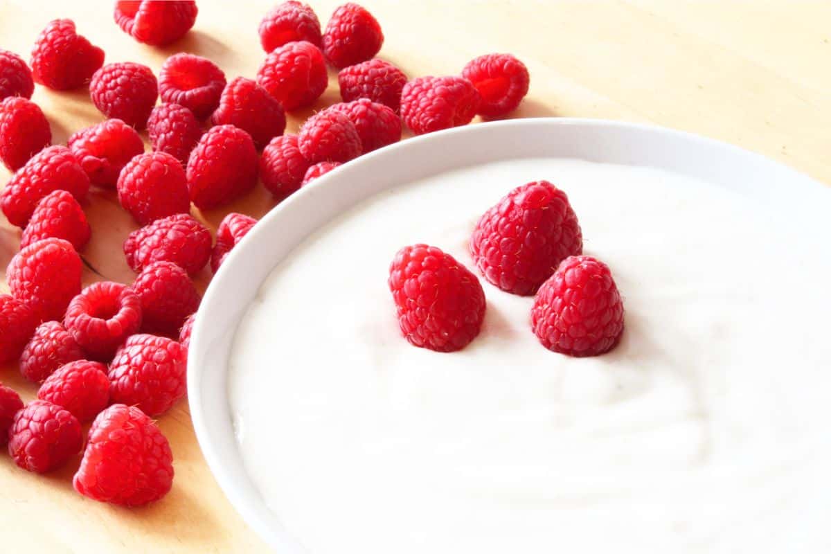 Bowl of plain plant-based yogurt topped with fresh raspberries on a light background.