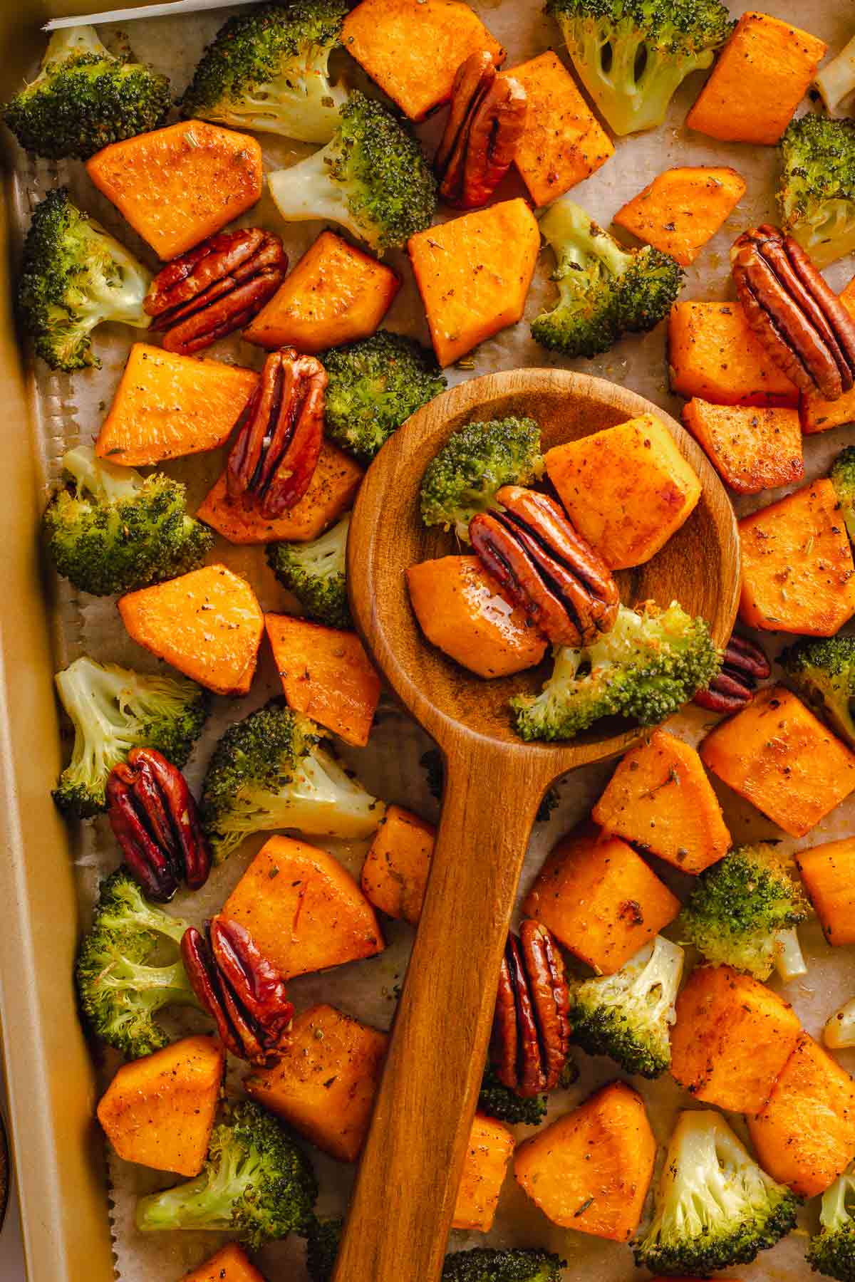 Wooden spoon lifting roasted broccoli and sweet potatoes from sheet pan
