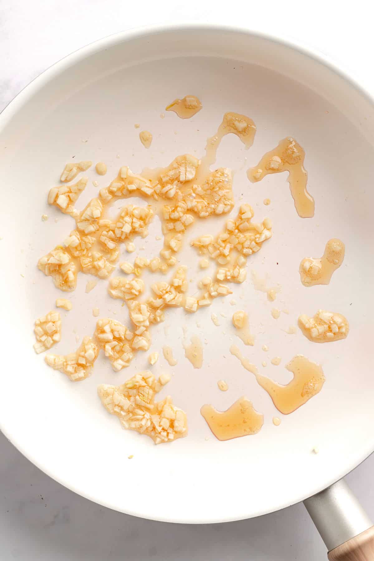 Minced garlic cooking in sesame oil in a white skillet.