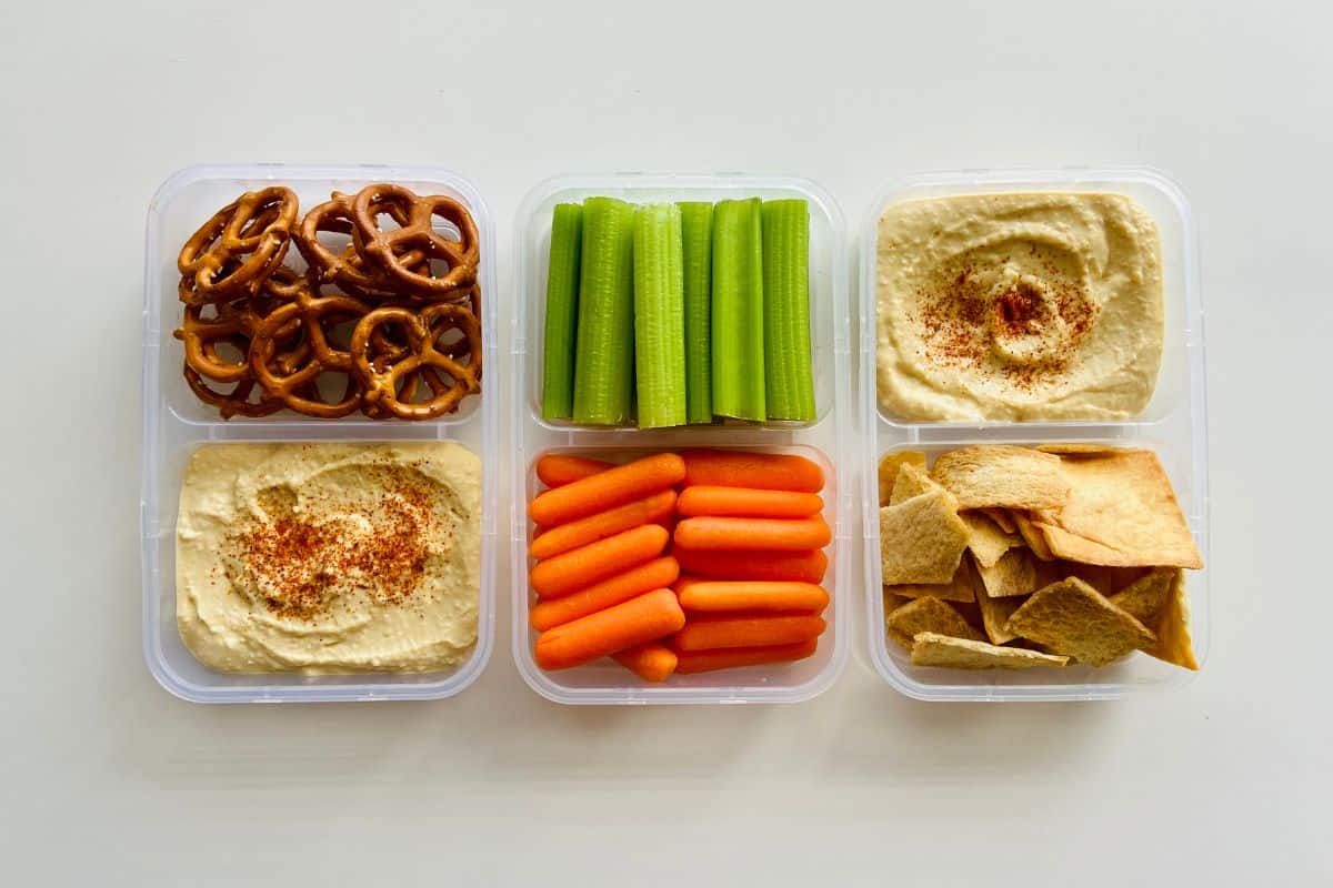 Vegan snack containers with hummus, vegetables, pretzels, and pita chips