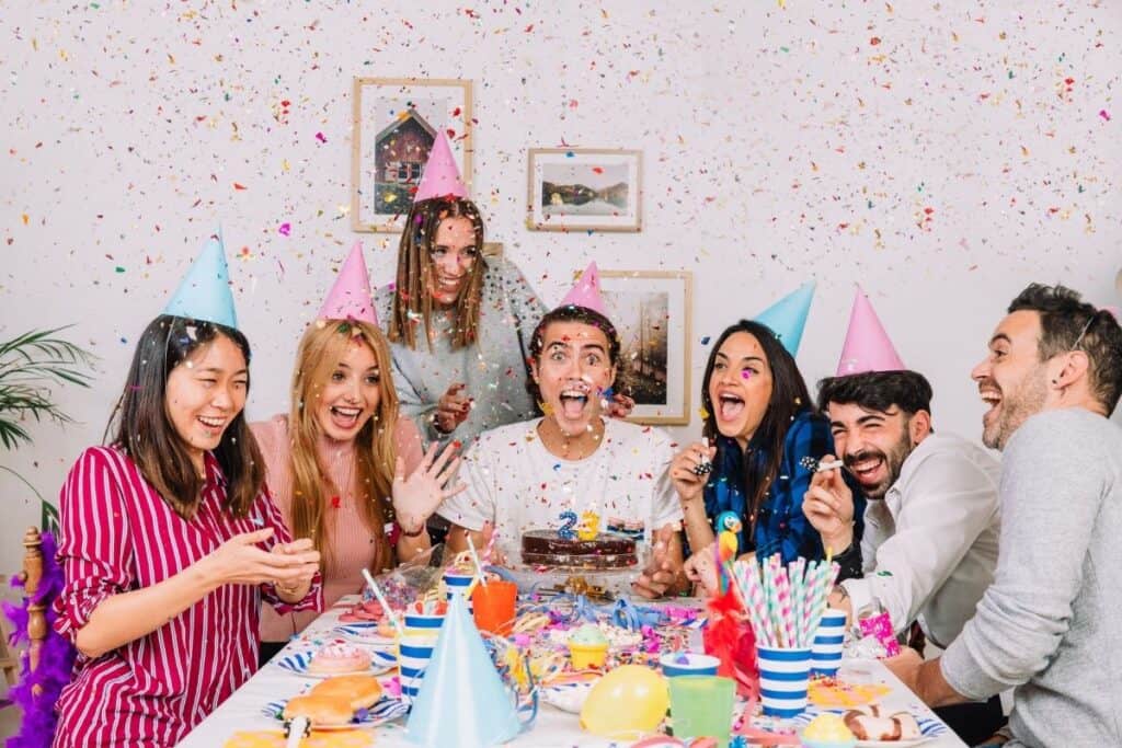 Group of friends celebrating a birthday together with party hats, confetti, food, and decorations.
