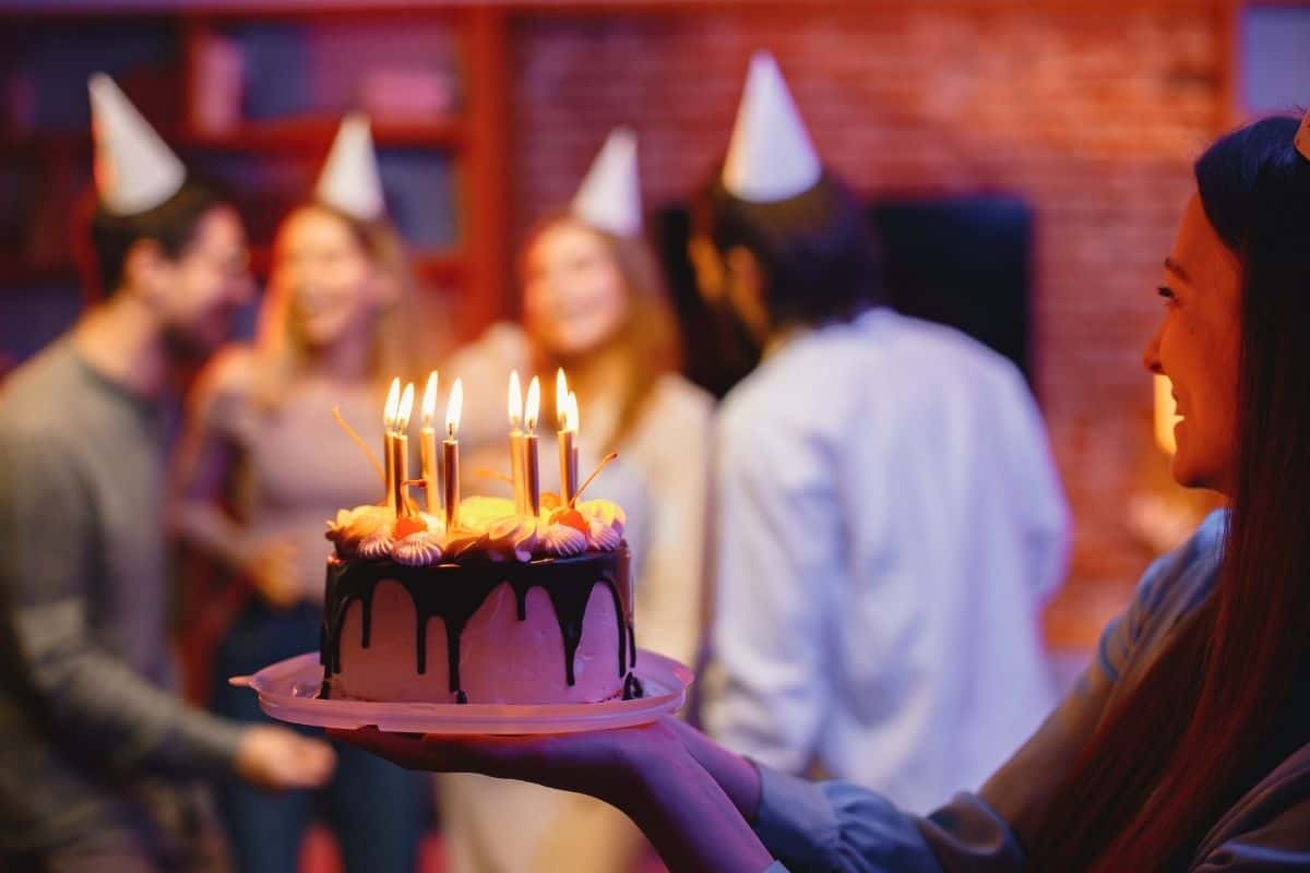 Woman carrying a birthday cake with lit candles toward a group of people at a party.