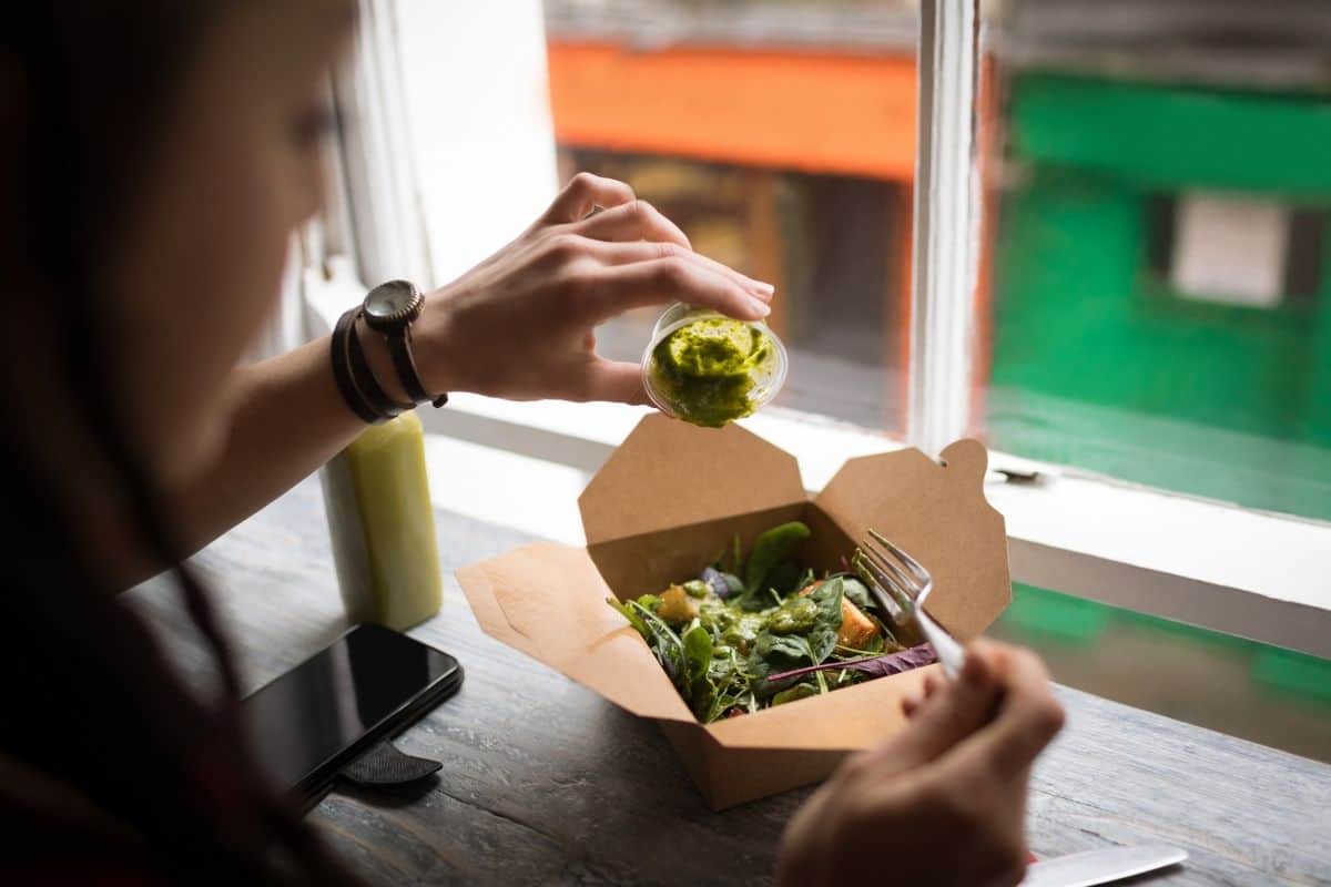Person adding dressing to a takeaway salad near a window.