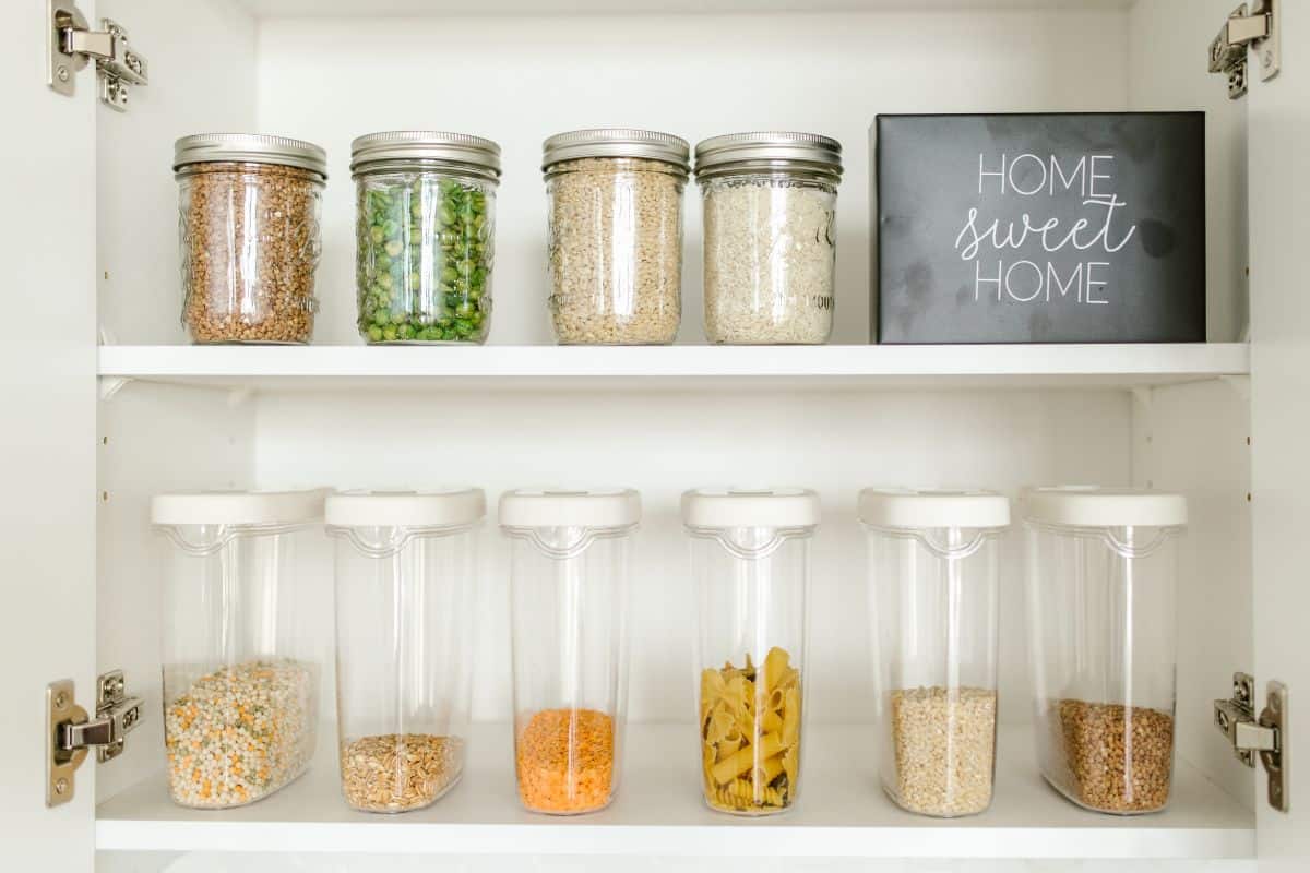 Glass jars of dry pantry ingredients arranged on white kitchen shelves.