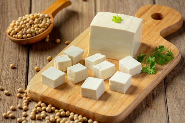 Cubes and a block of tofu on a wooden cutting board with soybeans and fresh herbs.