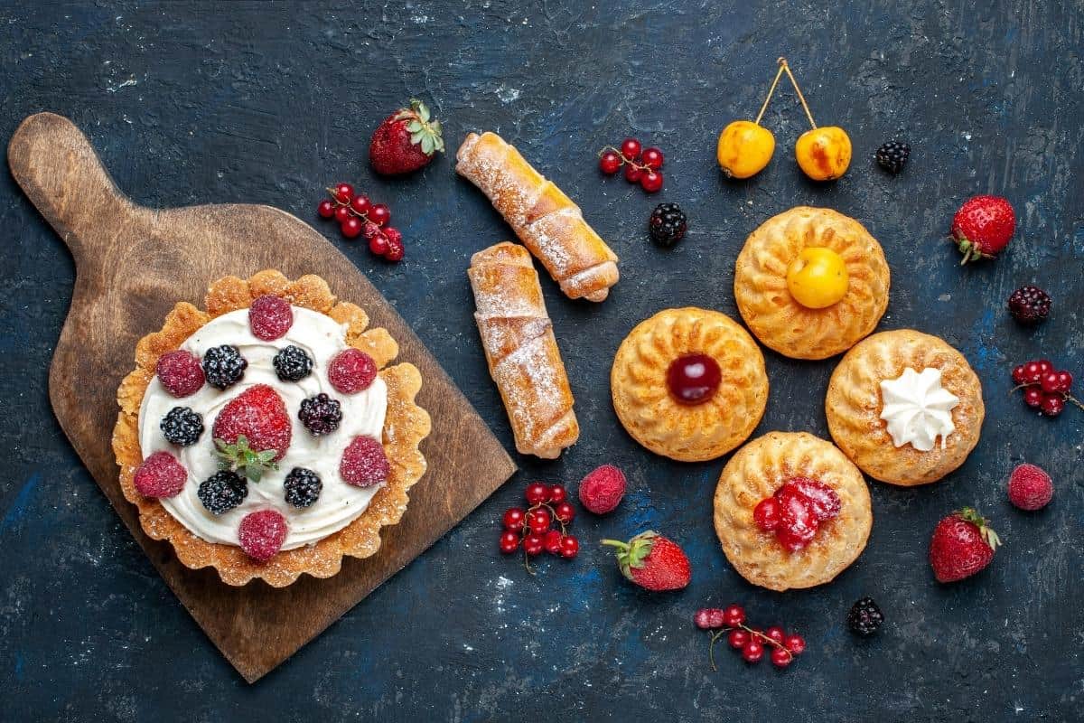 Assorted pastries and fruit tarts topped with berries on a dark surface.